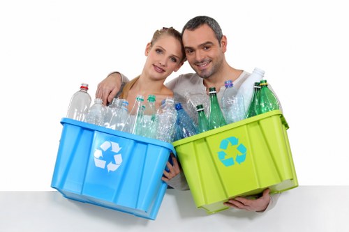 A smiling man and woman stand close together, each holding a plastic recycling bin, one blue and one green, filled with clear plastic bottles of varying shapes and sizes. The blue bin, held on the left, features a white recycling symbol on the side and contains mostly transparent bottles with some light green and clear hues, with visible ridges and caps. The green bin, on the right, also bears a white recycling symbol and is filled with similarly transparent plastic bottles, some with coloured caps. Both individuals are facing forward and are set against a plain white background, suggesting an indoor or studio setting. The scene emphasizes waste separation and recycling practices, consistent with rubbish removal and waste management services, highlighting environmentally conscious disposal of plastic materials often encountered during commercial waste collection in Cranford or surrounding areas.