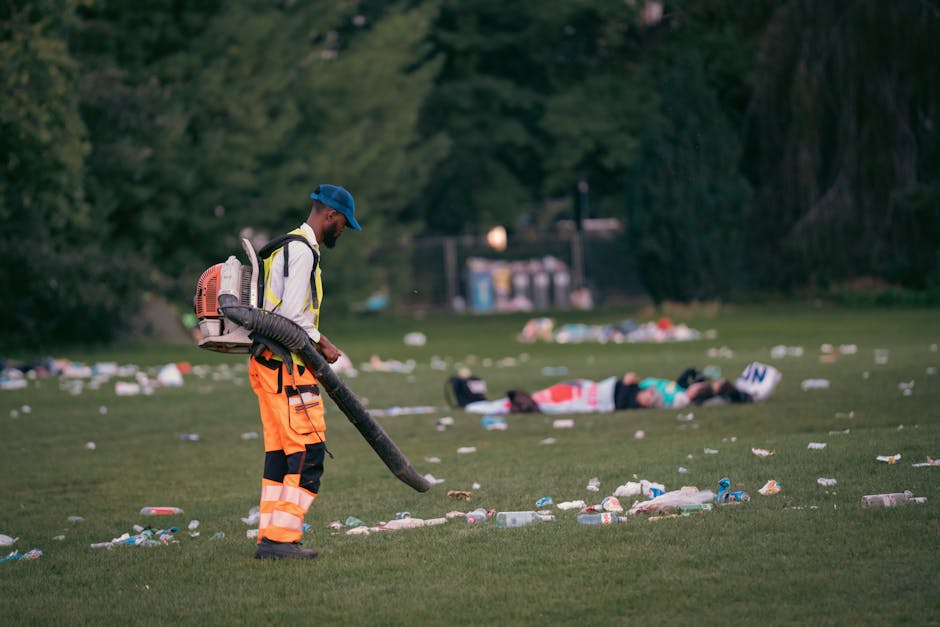 A worker dressed in high-visibility orange trousers with reflective stripes, a white shirt, a blue cap, and carrying a backpack blower, stands on a grassy park area during daylight hours. The worker i