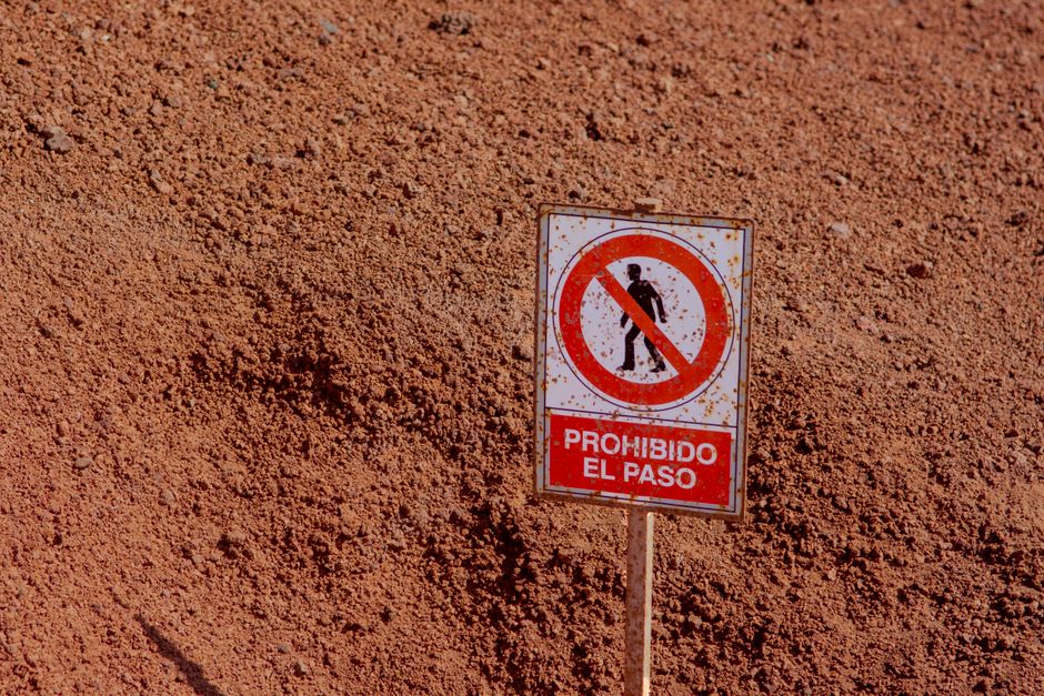 A rectangular 'No Trespassing' sign attached to a weathered wooden board, which is mounted on a rusted metal fence post. The sign has a black background with bold, uppercase, pink text that reads 'NO