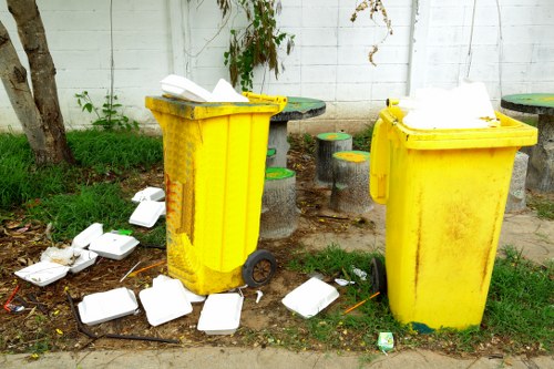 The image depicts two large, yellow plastic waste bins placed outdoors on a grassy area near a concrete wall and a tree trunk, with some dark patches on the ground. One bin is positioned on the left and the other slightly to the right, both with their lids open. Around the bins, several white foam takeout containers and small pieces of litter are scattered on the ground, some leaning against the bins and others lying directly on the pavement or grass. The background shows a white wall with visible brick patterns, partially obscured by a leafy green vine, and a few small stool-like objects painted in green, set behind the bins. The scene appears to be a disposal or rubbish collection point, with visible waste indicating recent generation or disposal of food packaging, typical of outdoor rubbish management practices. The overall setting suggests an urban or suburban environment, possibly in Cranford or a similar locality in the UK, where Commercial Waste Cranford offers rubbish removal services for such areas.