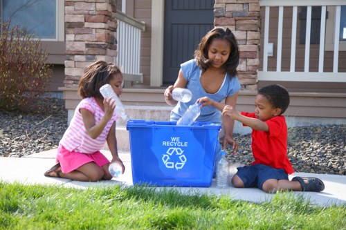 Three children, two girls and one boy, are engaged in recycling activities outdoors on a residential driveway in Cranford. The girl on the left, wearing a pink and white striped top and pink shorts, is holding a clear plastic bottle close to her mouth, appearing to prepare for recycling. The girl in the center, dressed in a blue top and denim shorts, is actively placing a plastic bottle into a blue recycling bin marked with the recycling symbol and the words 'WE RECYCLE.' The boy on the right, wearing a red shirt and navy shorts, is also holding a plastic bottle, assisting in the recycling process. The background features a modern house with a brick and wood facade, a dark gate, and a porch railing, with a well-kept garden area with gravel and small plants. The scene is illuminated by natural daylight, emphasizing the clean, fresh environment and the children's focus on environmentally conscious activity, reflecting waste management practices suitable for residential areas near Cranford.