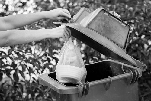 A person is disposing of waste into a black wheelie bin positioned outdoors on a paved area, with the lid open. The individual is placing a clear plastic bag containing white dishware or bowls into the bin, holding it steady with both hands. The bin is situated near a backdrop of dense, leafy foliage, suggesting a garden or driveway setting in Cranford. The bin's surface appears to be slightly textured and weathered, with visible hinges and handles. The scene is lit with natural daylight, illuminating the waste disposal activity. This image illustrates proper rubbish disposal practices, relevant to commercial waste management services by Commercial Waste Cranford, highlighting the handling of household or light waste materials as part of routine rubbish collection in the local area.